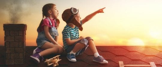 Two kids sitting on the roof of a house pointing at the sunny sky